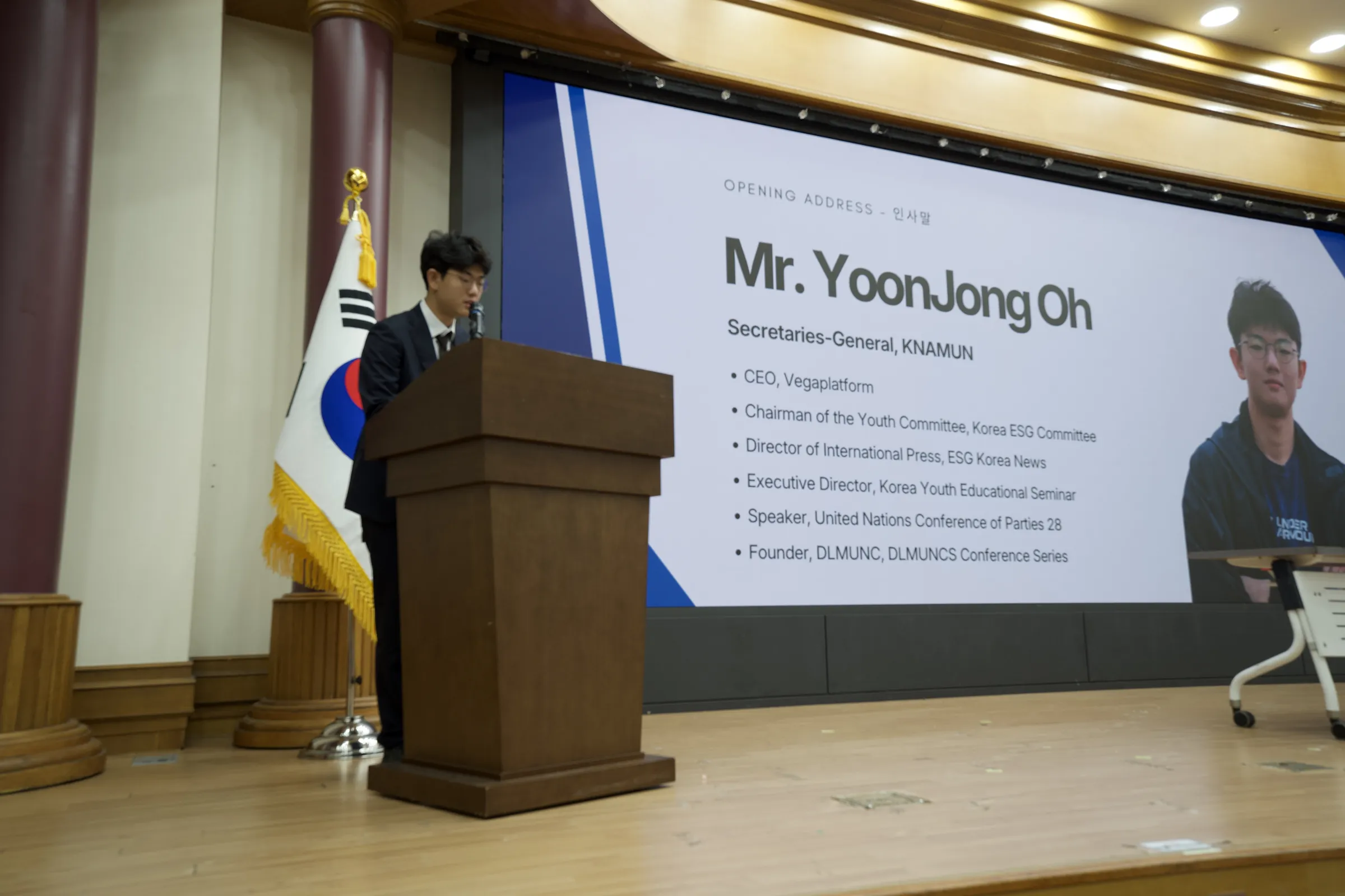 A speaker in a dark suit stands at a wooden podium beside a Korean flag, in front of a large screen titled Opening Address, Mr. YoonJong Oh, Secretary-General, KNAMUN.
