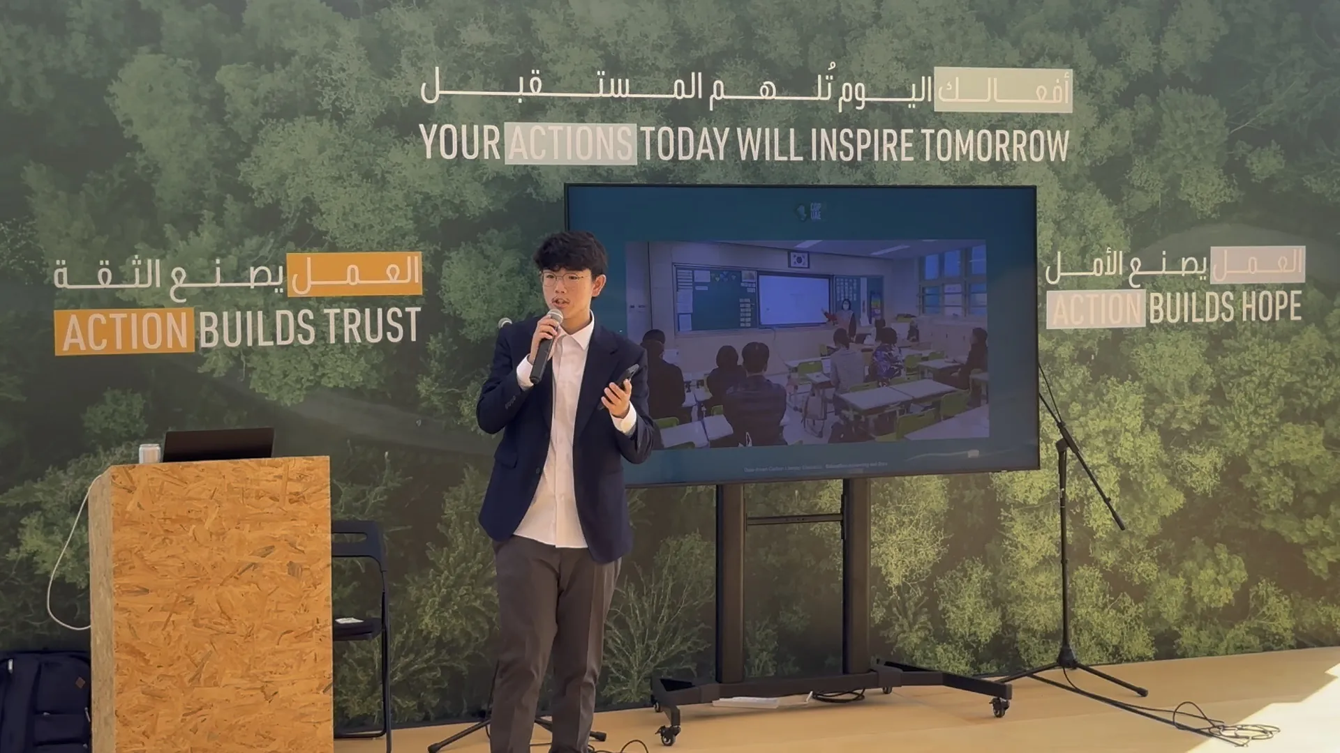 A young man in a navy blazer speaks into a handheld microphone, phone in his other hand, in front of a screen showing a classroom and a forest-print wall reading Your actions today will inspire tomorrow, Action builds trust, Action builds hope.