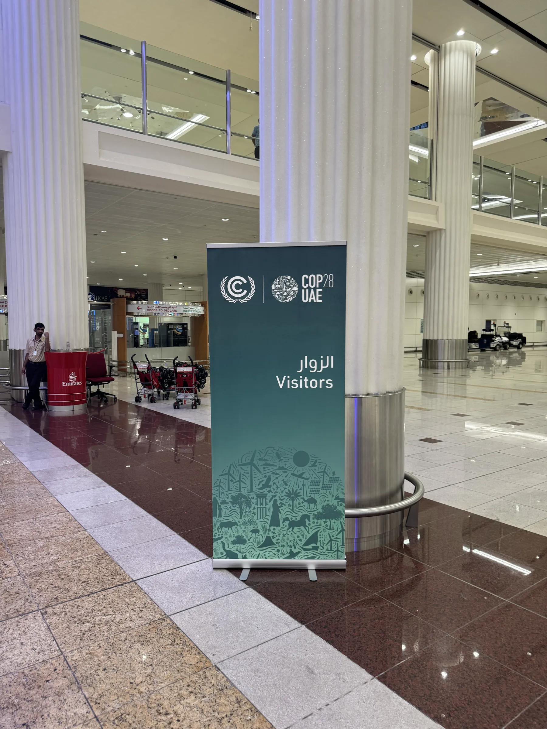 A teal COP28 UAE banner marked Visitors stands between marble columns in a quiet airport arrivals hall.