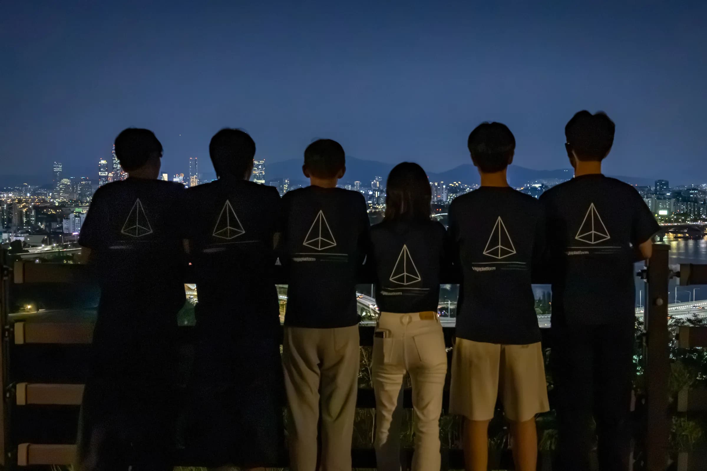 Six team members in matching black Vegaplatform T-shirts, seen from behind, look out over the lights of Seoul at night.